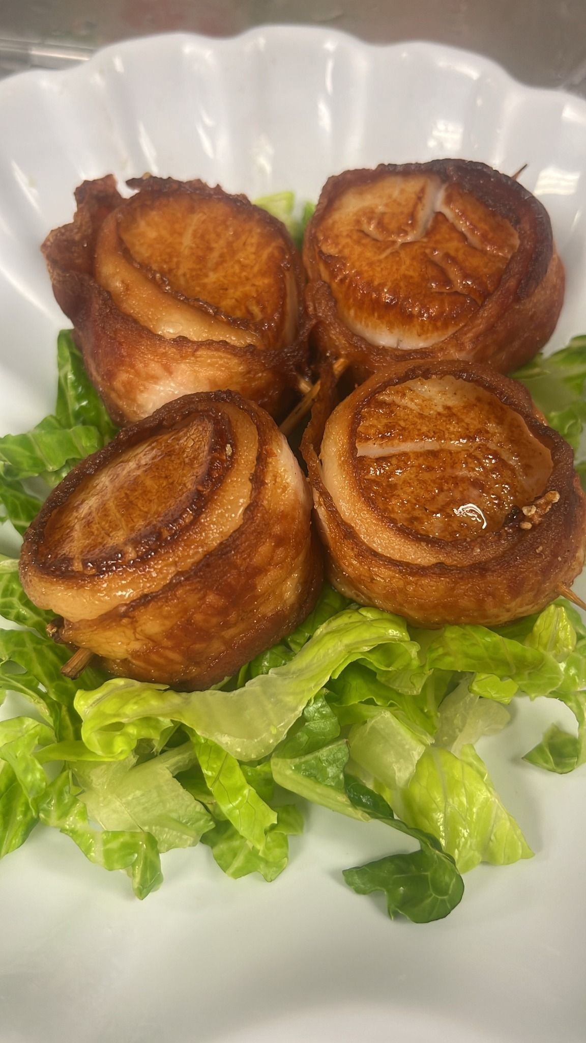 Four bacon-wrapped scallops on a bed of lettuce, arranged on a white scalloped plate. At High Tide Harry's