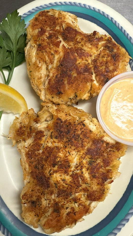 Two crab cakes on a plate with dipping sauce, lemon, and parsley. At High Tide Harry's