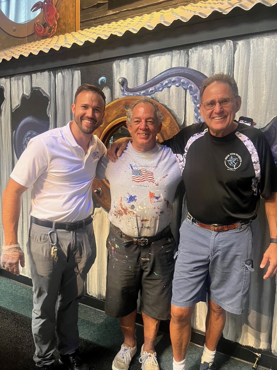 Three men pose in front of a painted wall. Two have arms around the middle man's shoulders, smiling.