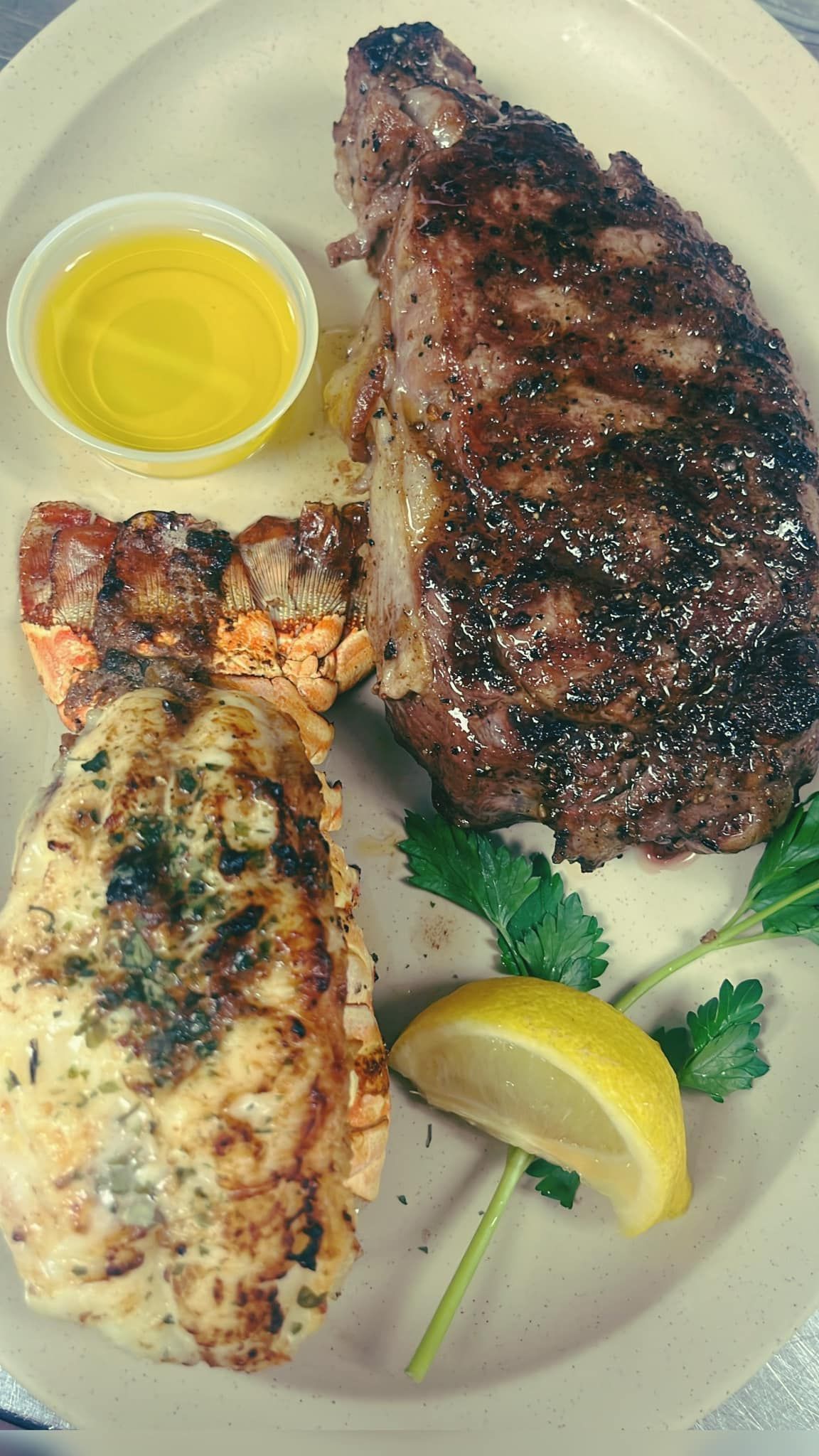 Steak and lobster tail with a cup of butter, lemon slice, and parsley on a plate at High tide Harry's