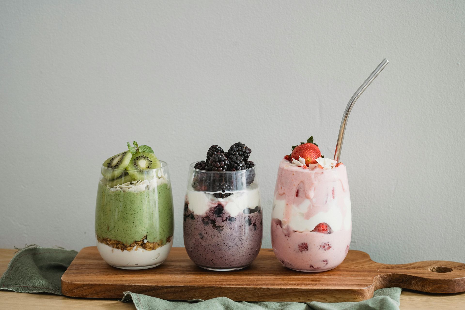 Three layered fruit smoothies in glass cups on a wooden board: kiwi, blackberry, and strawberry.