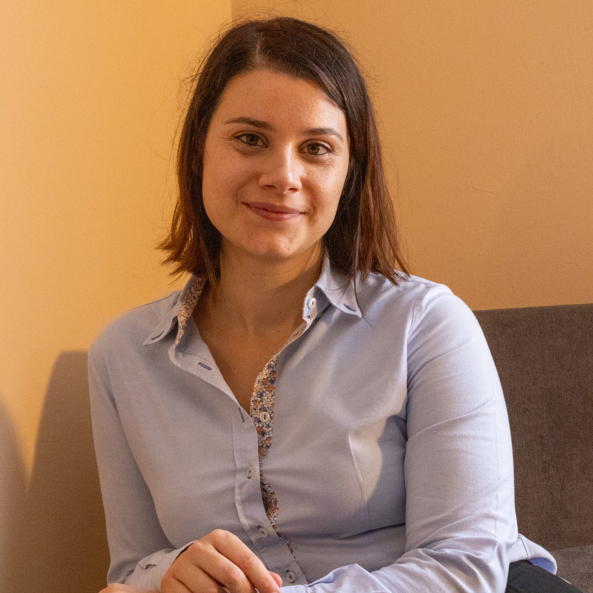 Una donna con i capelli castani, che indossa una maglietta azzurra, sorride alla telecamera, in un interno.