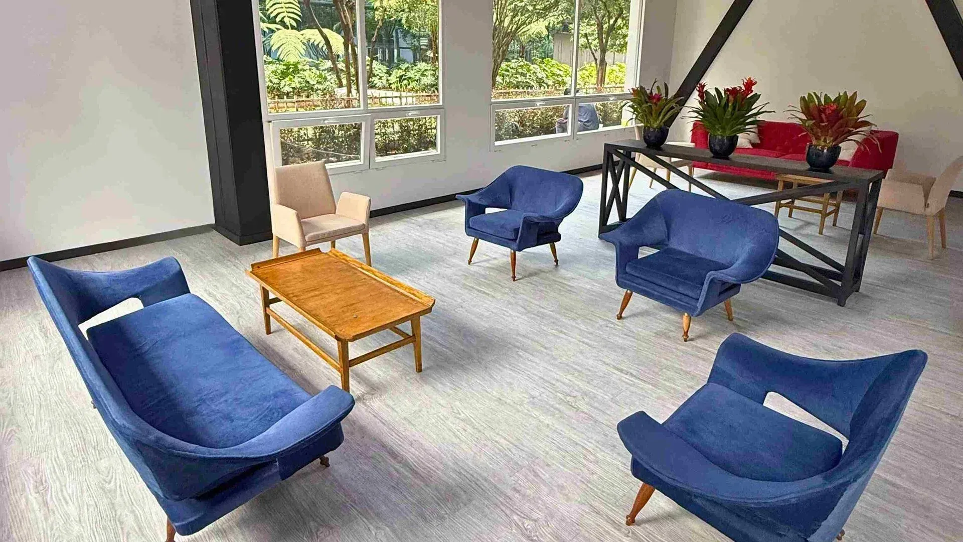 Poltronas azuis e uma mesa de madeira em uma sala iluminada com grandes janelas e arranjos de flores.