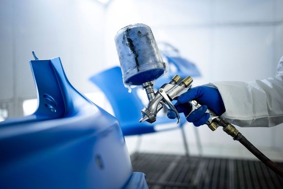 A person in protective gear sprays blue paint on a car part inside a paint booth. A person in protective gear sprays blue paint on a car part inside a paint booth.