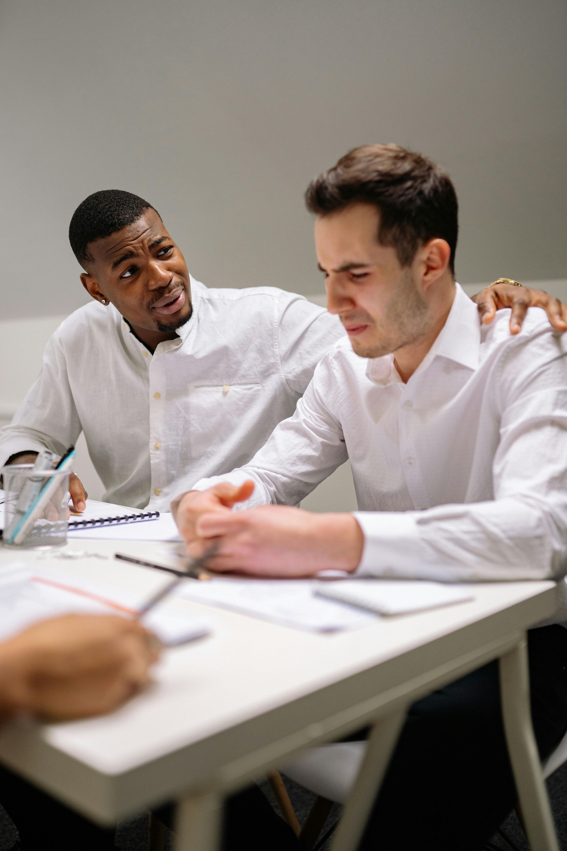 Two coworkers in white shirts reviewing documents at a desk, one consoling the other in a meeting room.