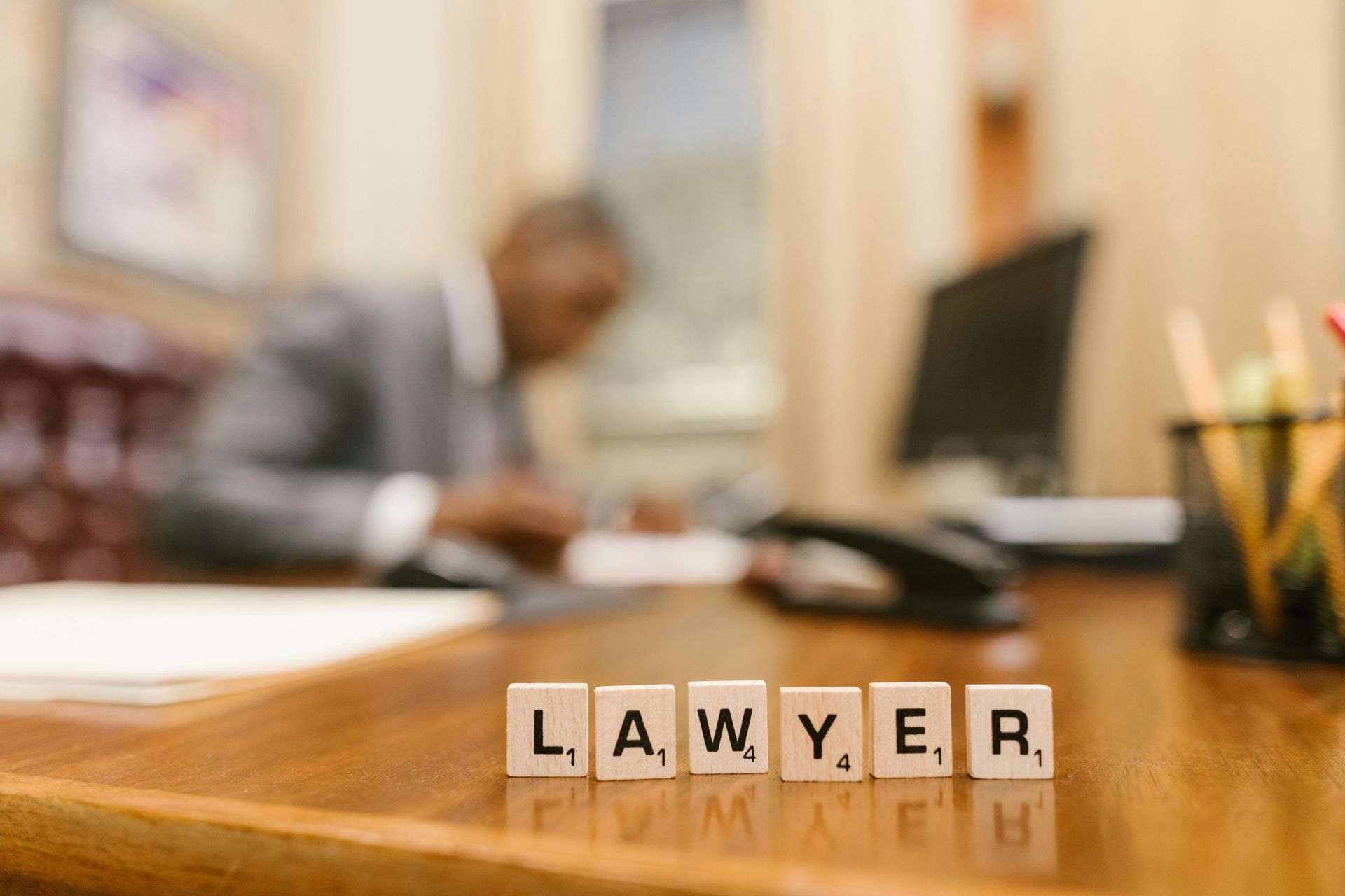 Scrabble tiles spelling “LAWYER” on a desk with a blurred office scene and person in background