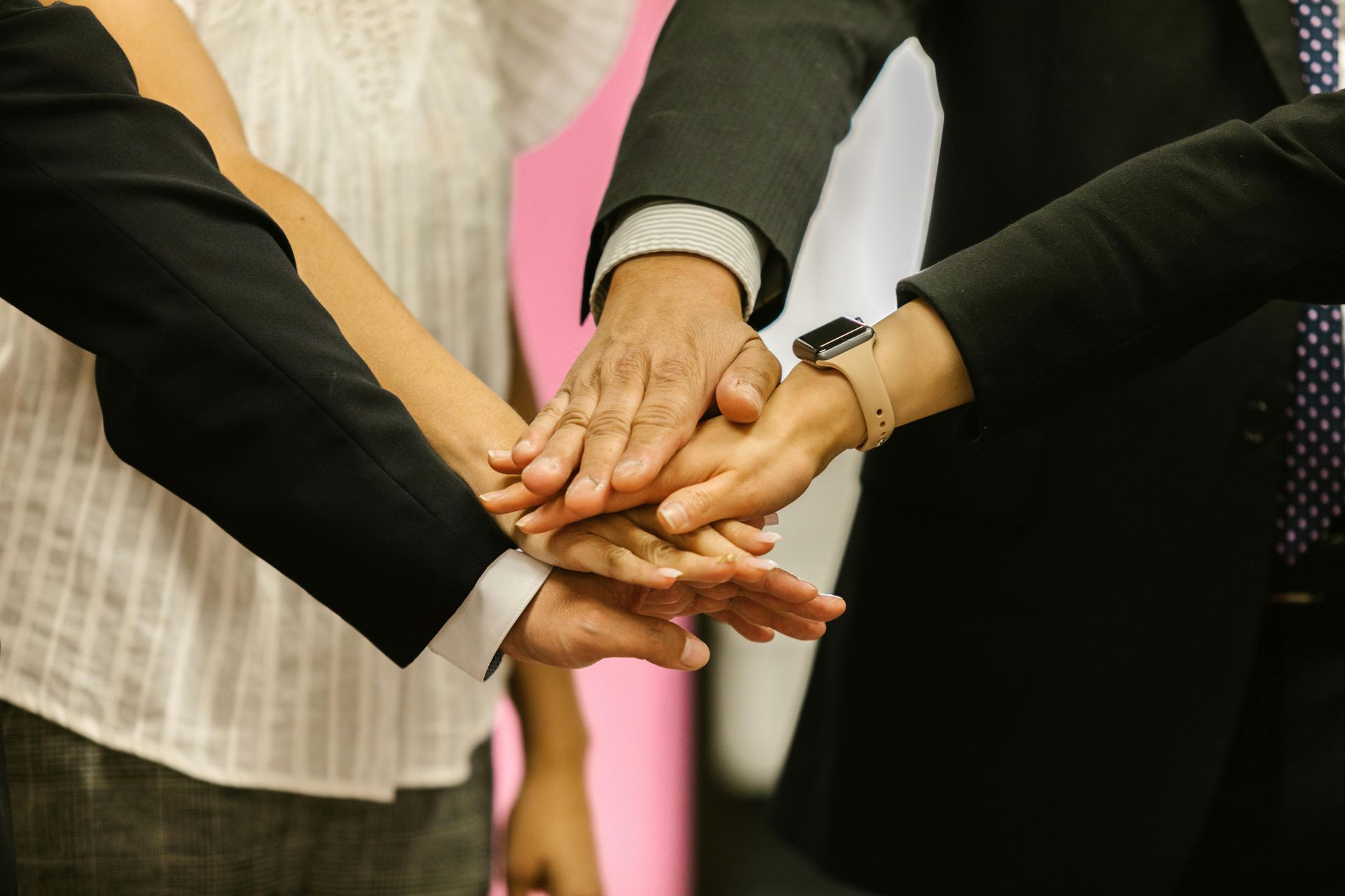 Hands stacked together in a team huddle, with suits and a pink background.