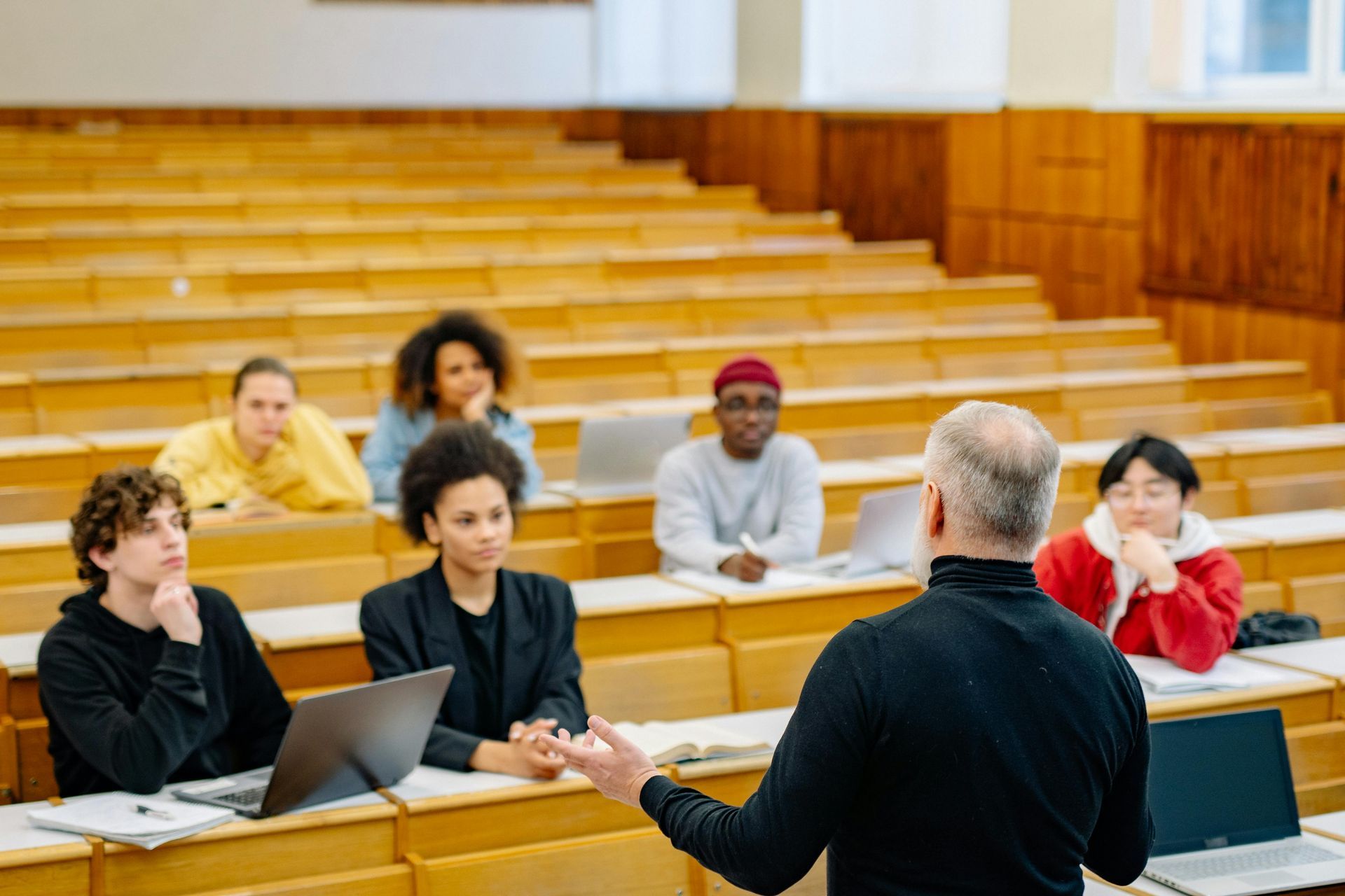 Instructor speaking to students in a lecture hall with wooden benches and laptops open.