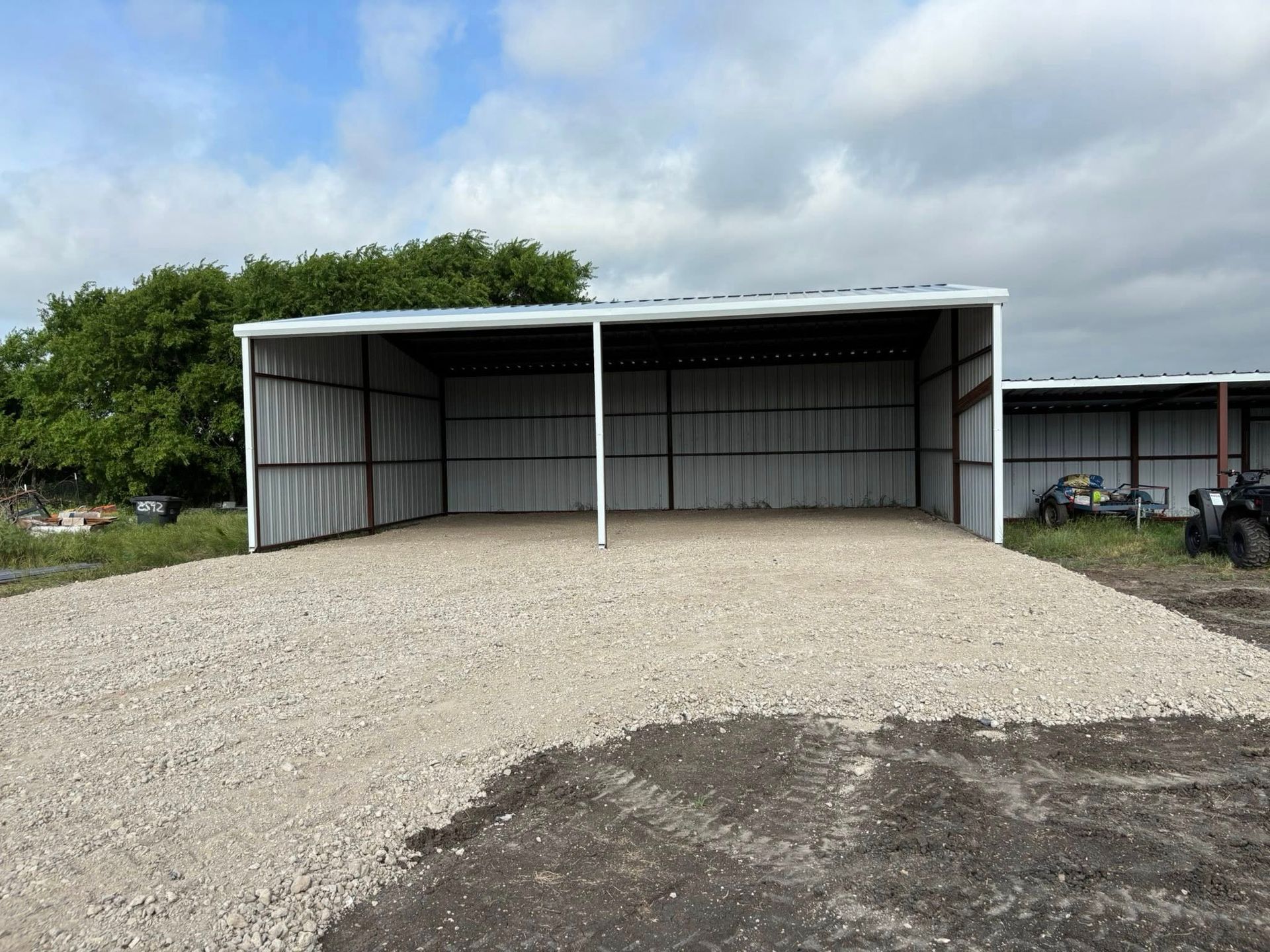 Open-sided metal shed with gravel flooring, against a cloudy sky.