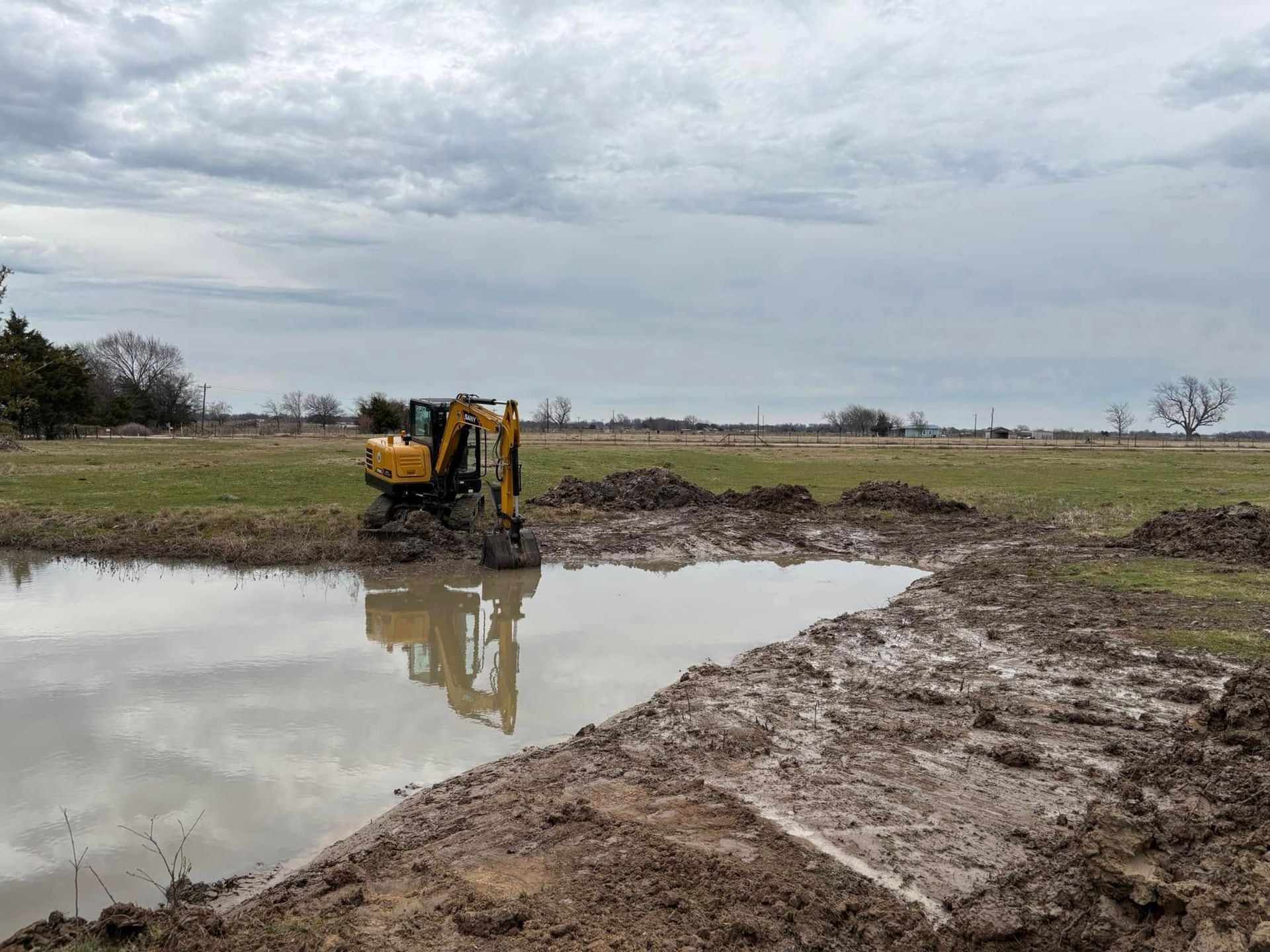 Excavator working in a muddy pond, digging and moving dirt in a field under cloudy skies.
