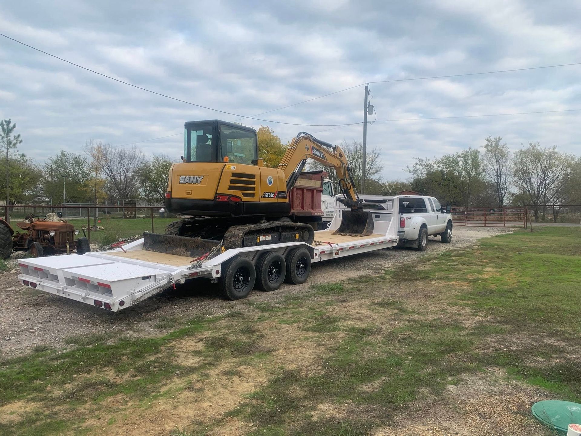 Yellow excavator on a trailer being pulled by a white truck on a gravel driveway under a cloudy sky.