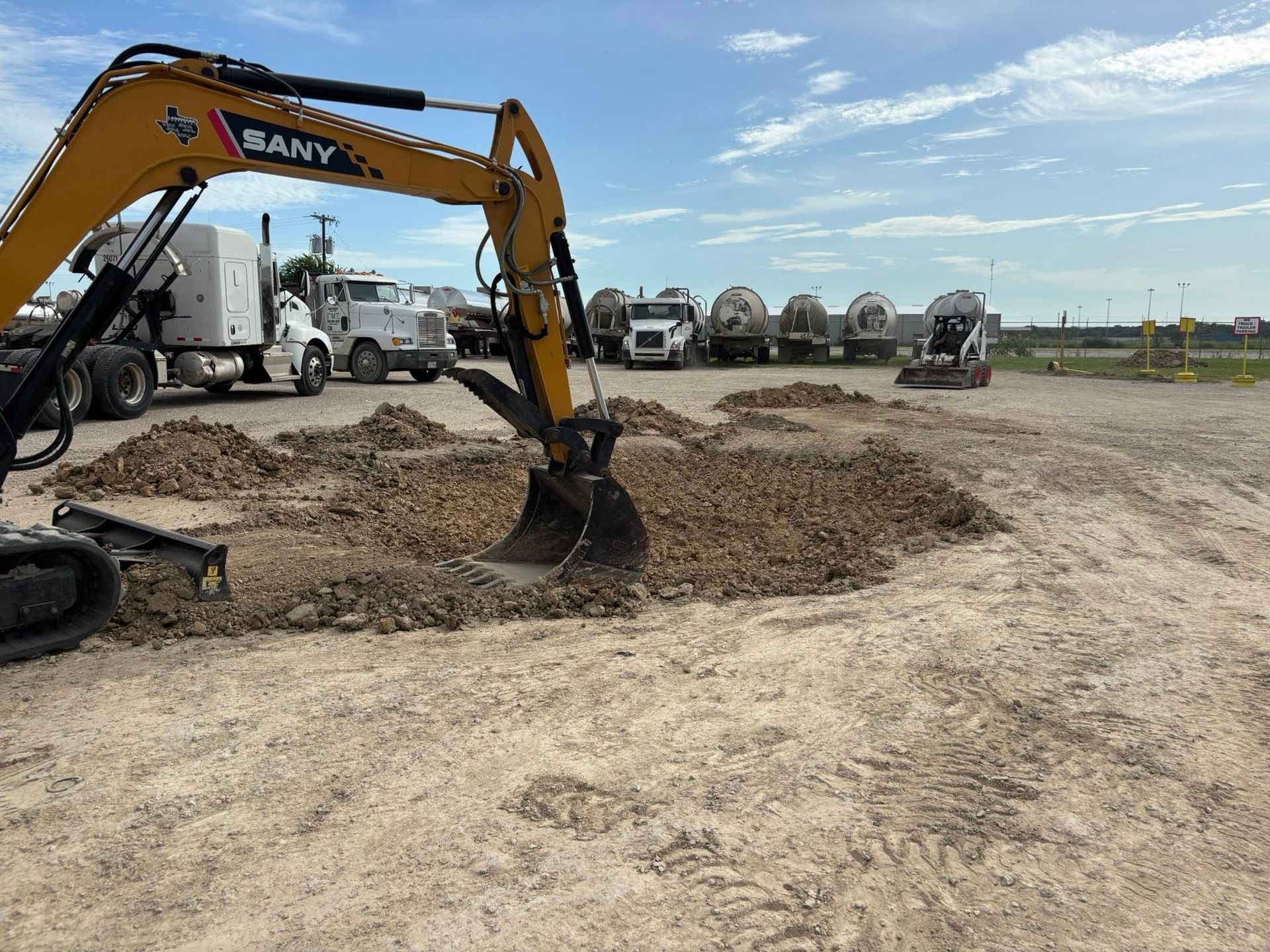 Yellow excavator digging in dirt lot; trucks in background.