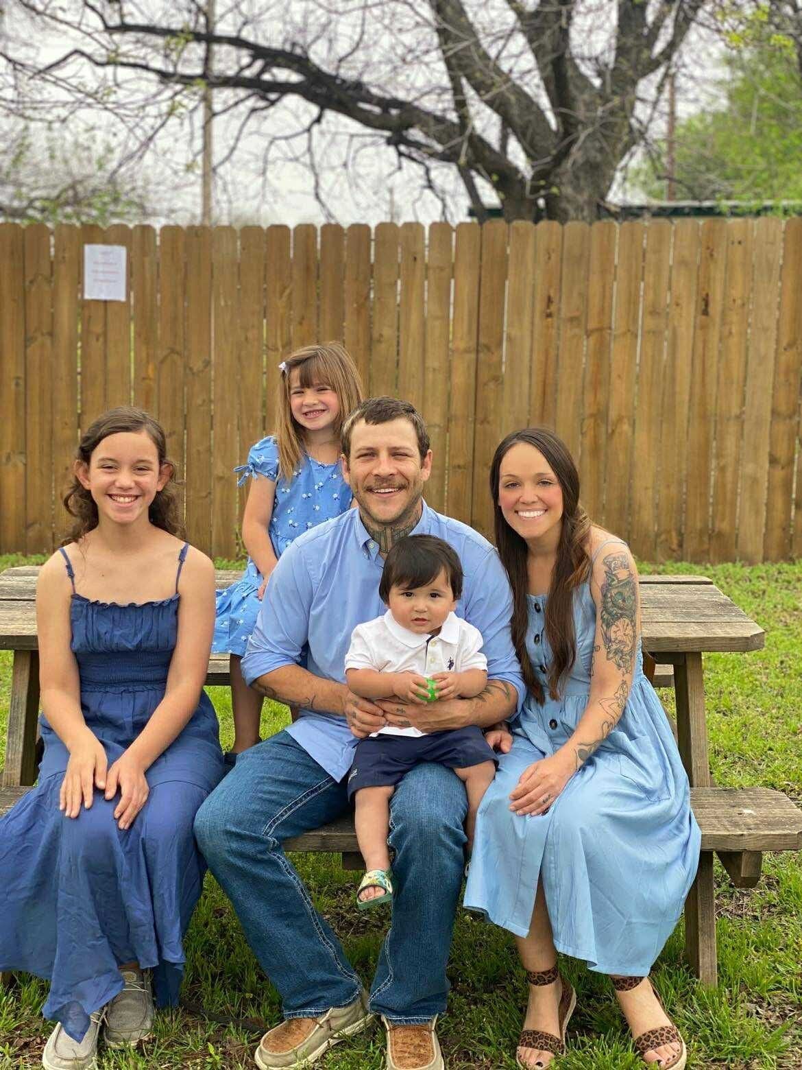 Family of five sitting on a wooden bench outdoors; all in blue, smiling at the camera, with a wooden fence background.