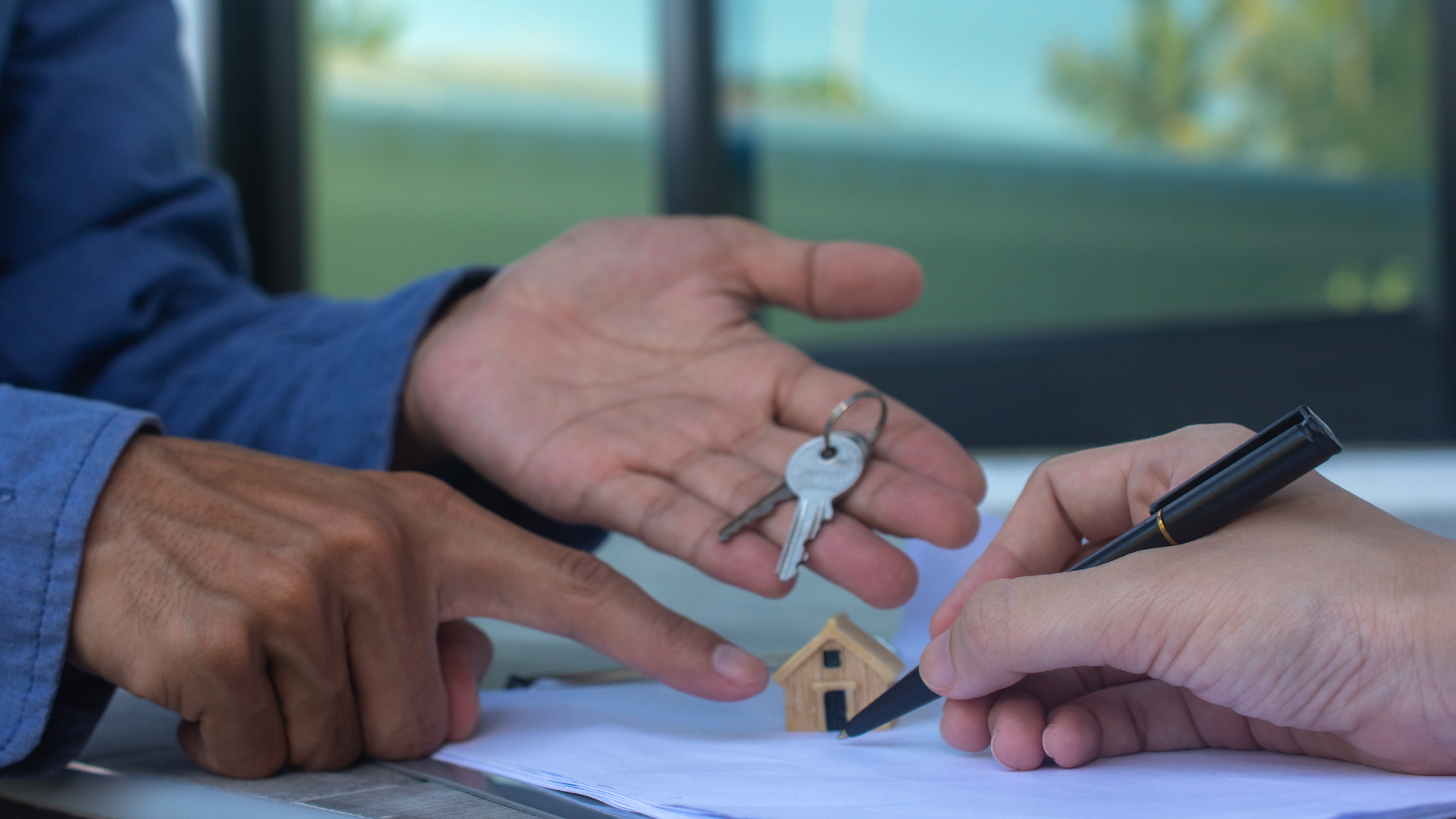 A hand holds keys over a small wooden house model while another hand signs a document, symbolizing a real estate deal.