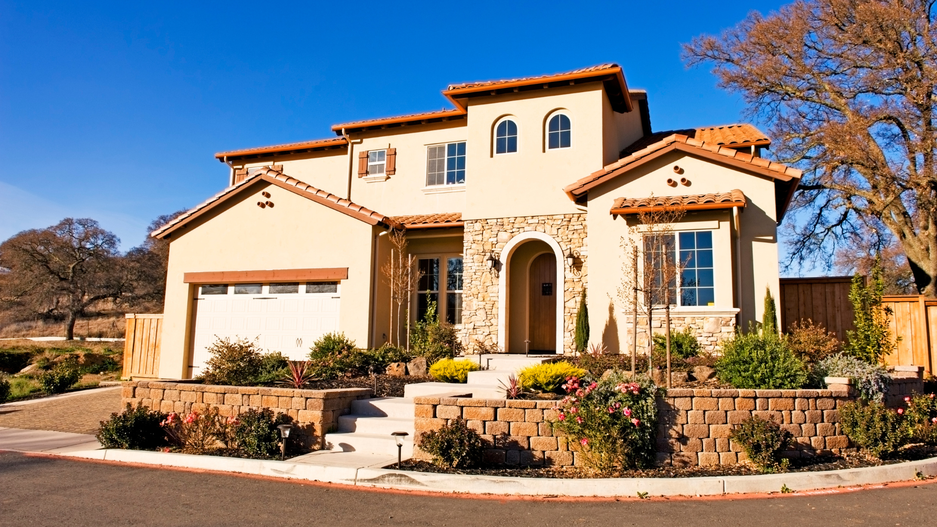 A two-story stucco suburban house with a stone entryway, arched front door, and a retaining wall under a clear blue sky.