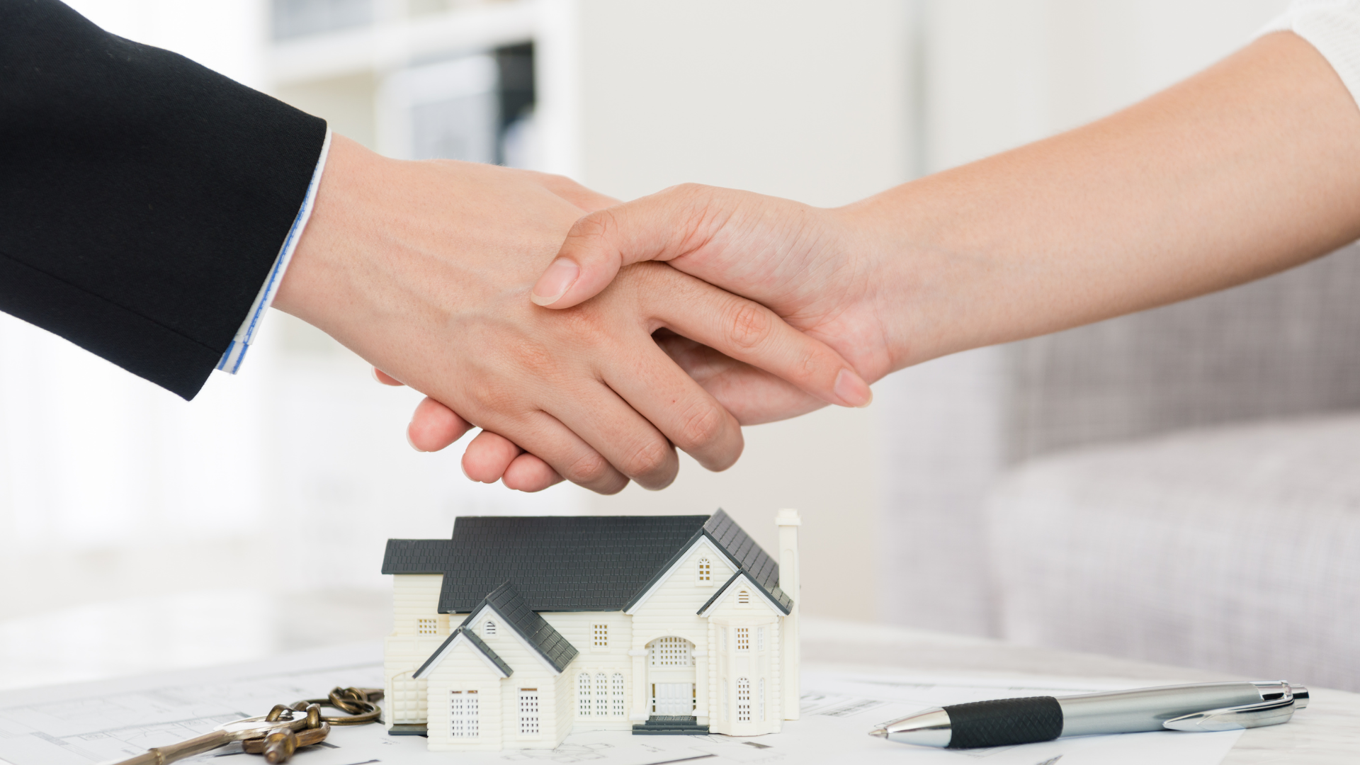 Two people shaking hands over a miniature house model, keys, and a pen on a table, symbolizing a real estate deal.