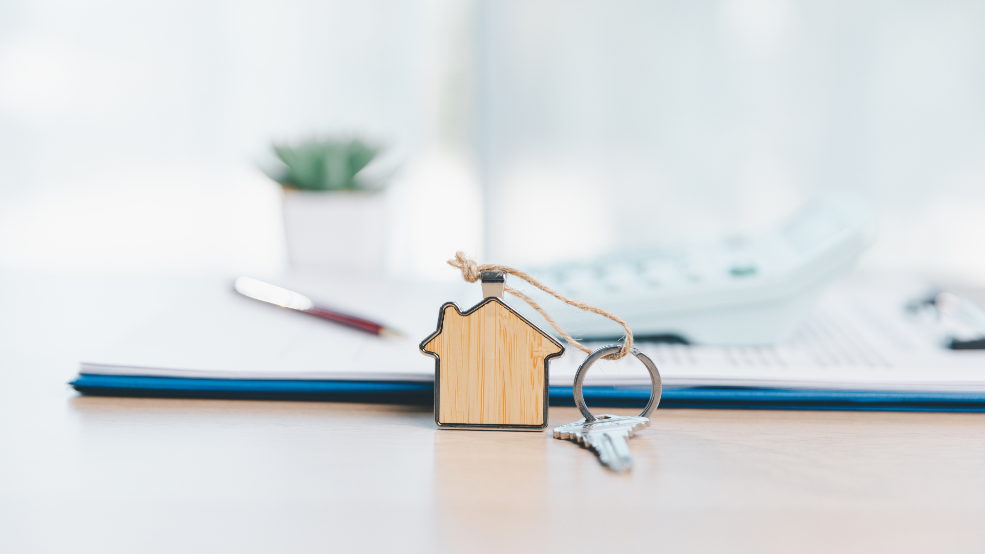 A wooden house-shaped keychain with a silver key resting on a document, with a calculator and small plant in the back.