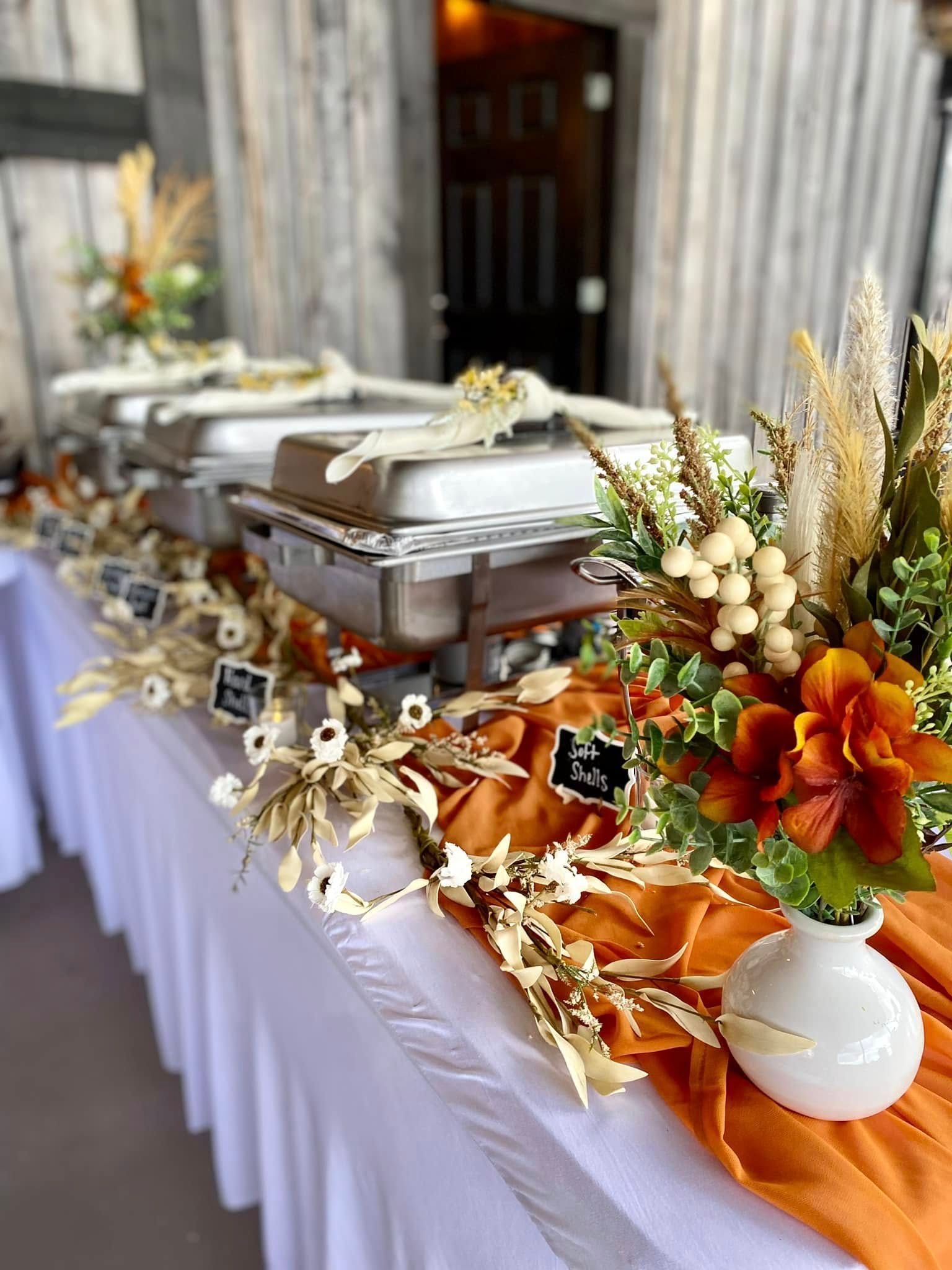 A long table filled with food in catering pans and beautiful decorations in orange and cream colors.