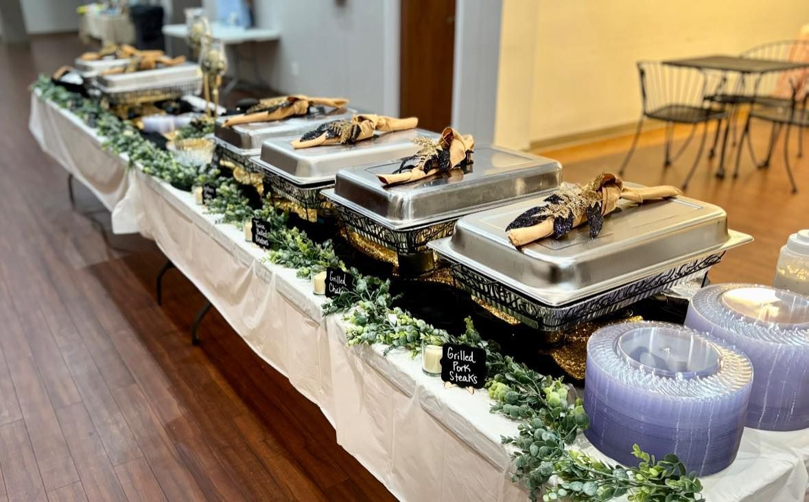 A long table with trays of food and plastic plates buffet style