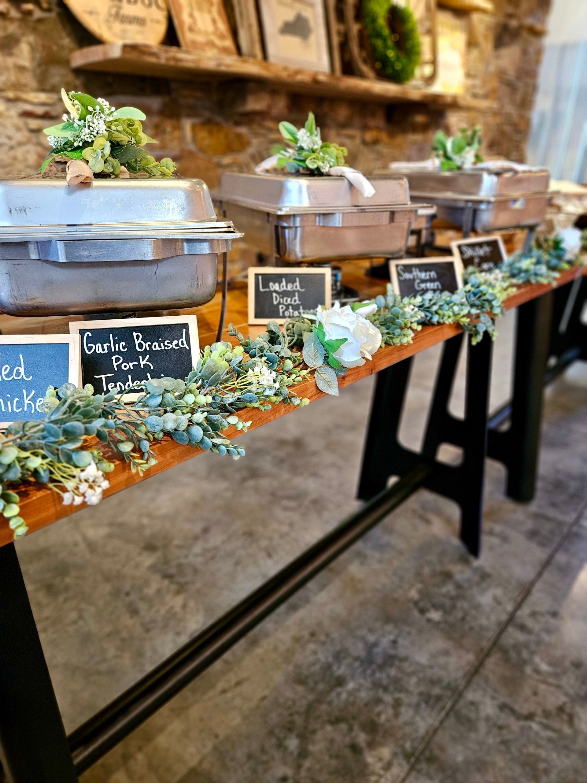 A wooden table with a bunch of covered trays of food on it with littel chalkboards noting the contents