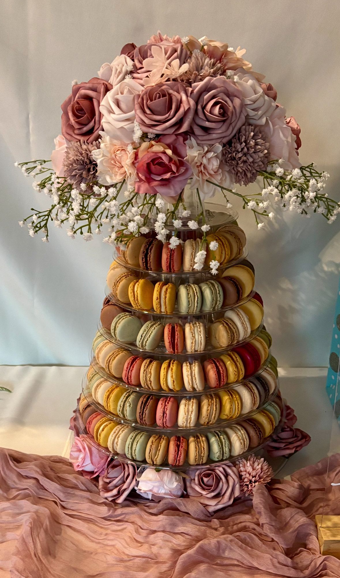 A stack of macarons sitting on a table, topped with a vase of roses in various shades of pink with white baby's breath.
