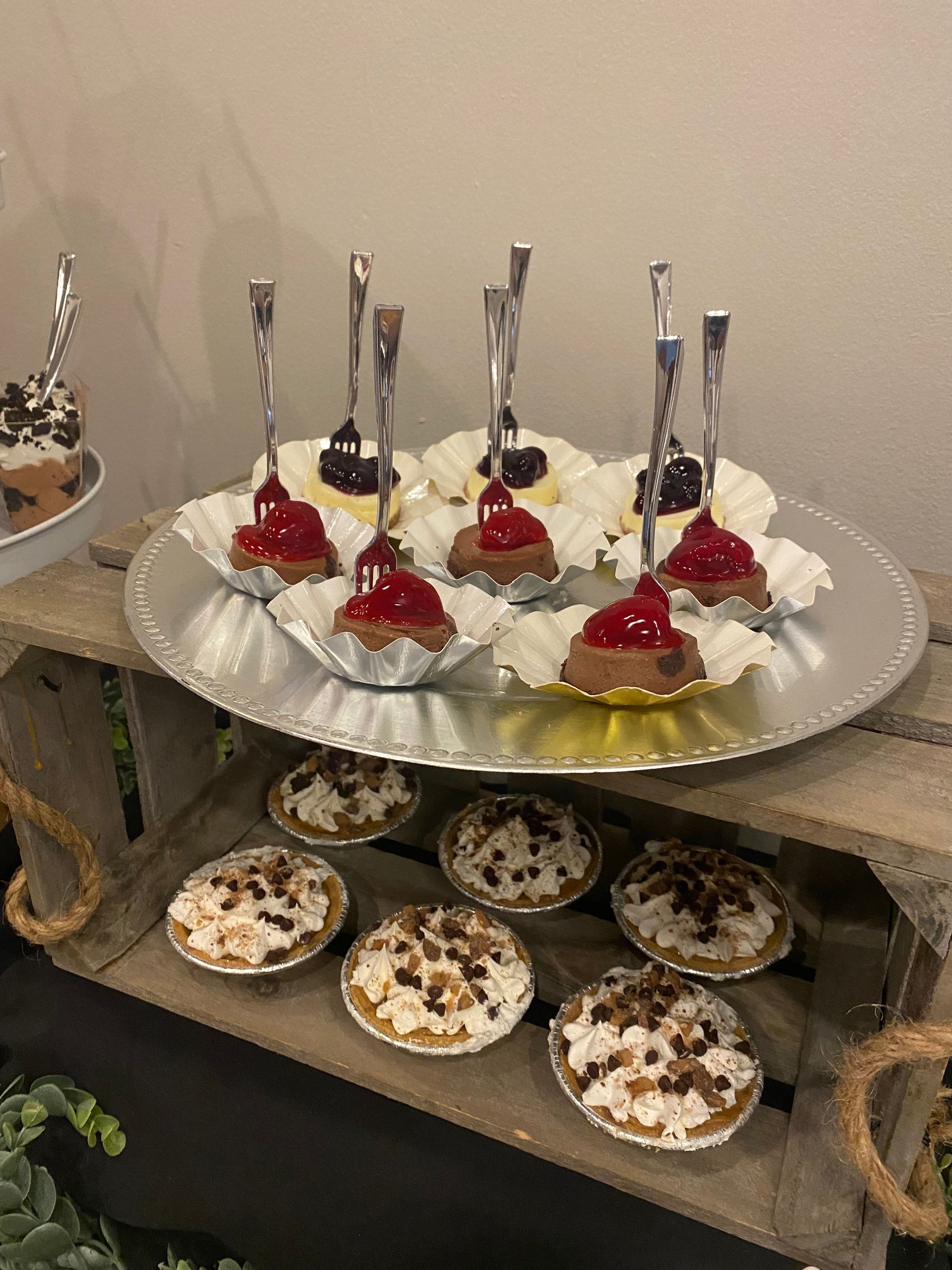 A silver plate topped with mini cheesecake rounds, topped with cherry or blueberry filling, and pies on a wooden table.