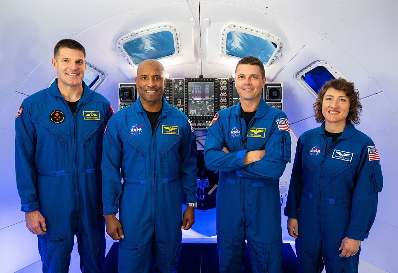 Four astronauts in blue flight suits standing in front of a spacecraft display