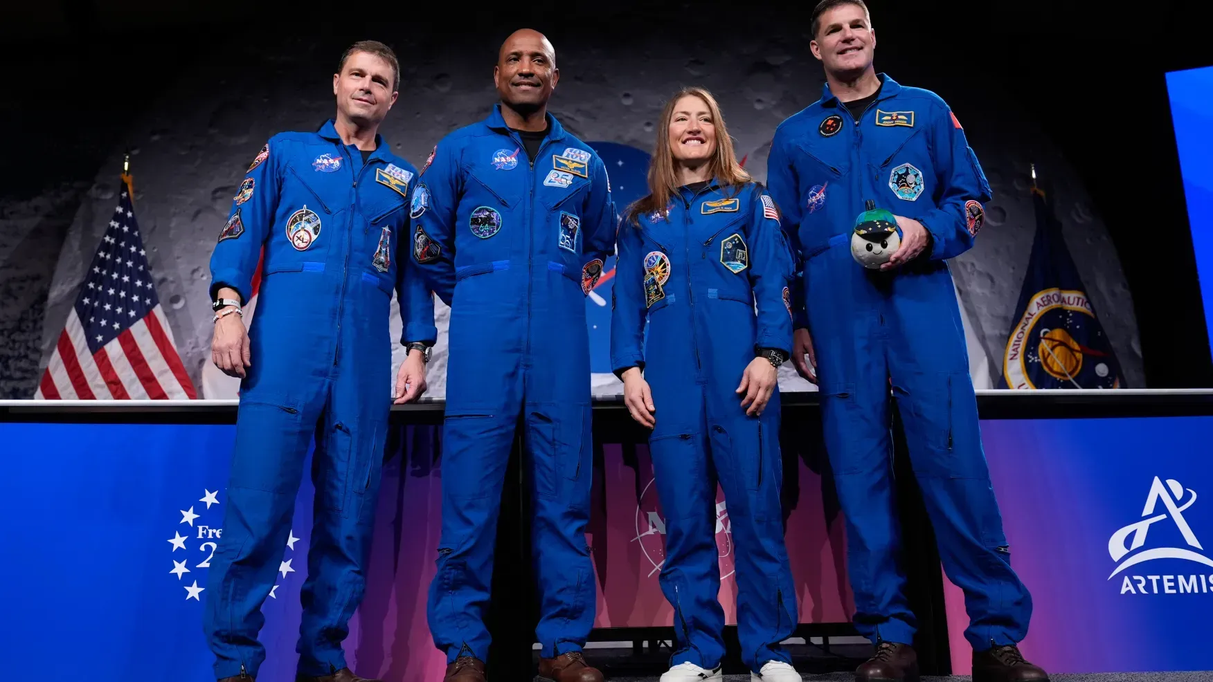 Four astronauts in blue flight suits stand onstage in front of U.S. and NASA flags