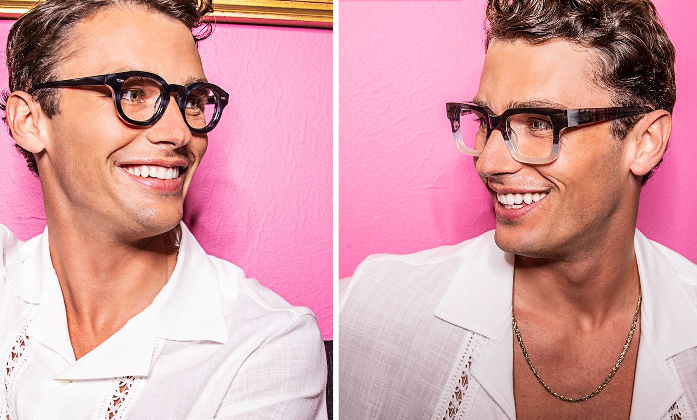 A man wearing glasses and a white shirt is smiling in front of a pink wall.