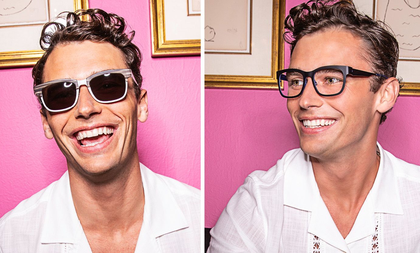 A man wearing sunglasses and glasses is smiling in front of a pink wall.