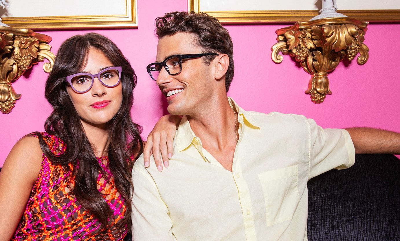 A man and a woman wearing glasses are sitting next to each other in front of a pink wall.