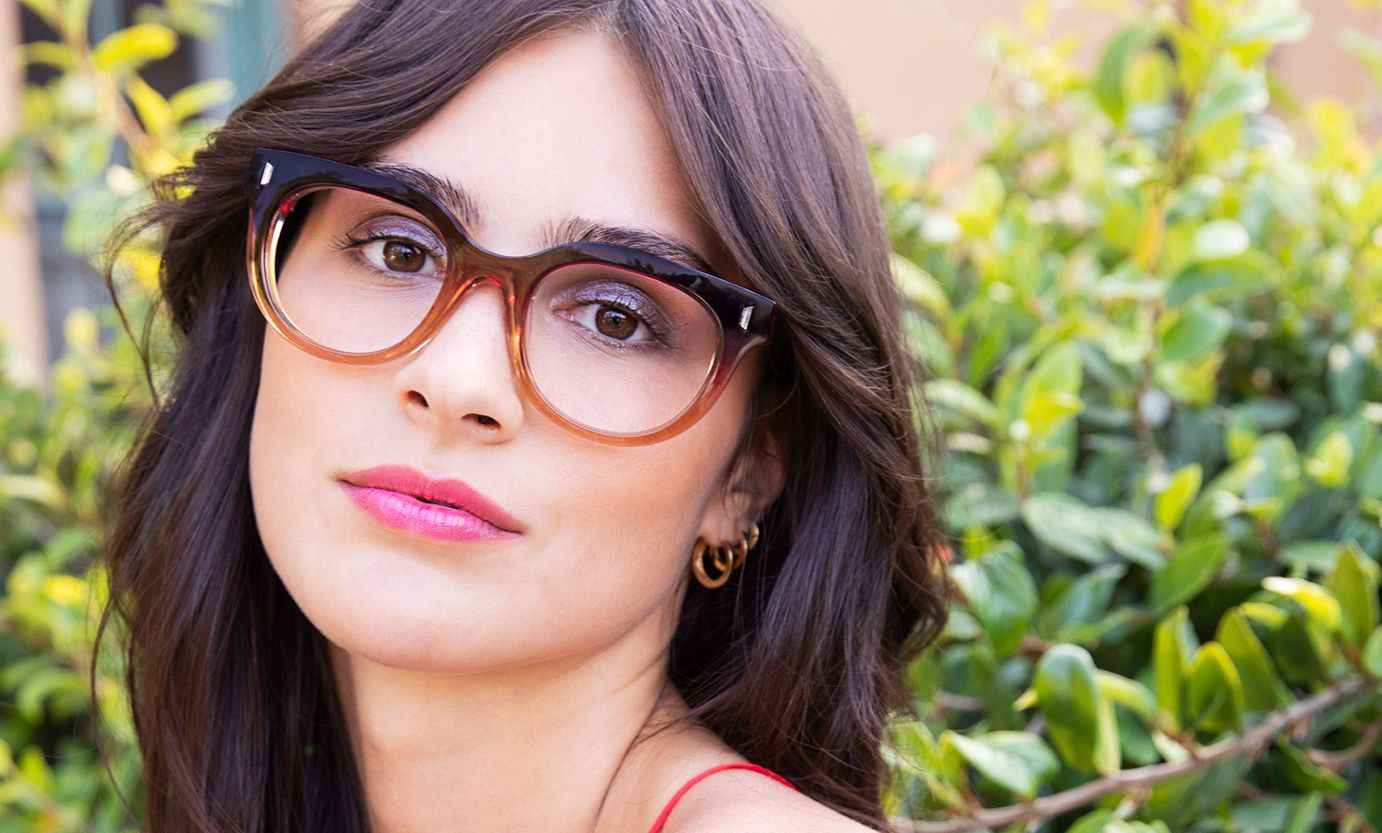 A woman wearing glasses and earrings is standing in front of a bush.