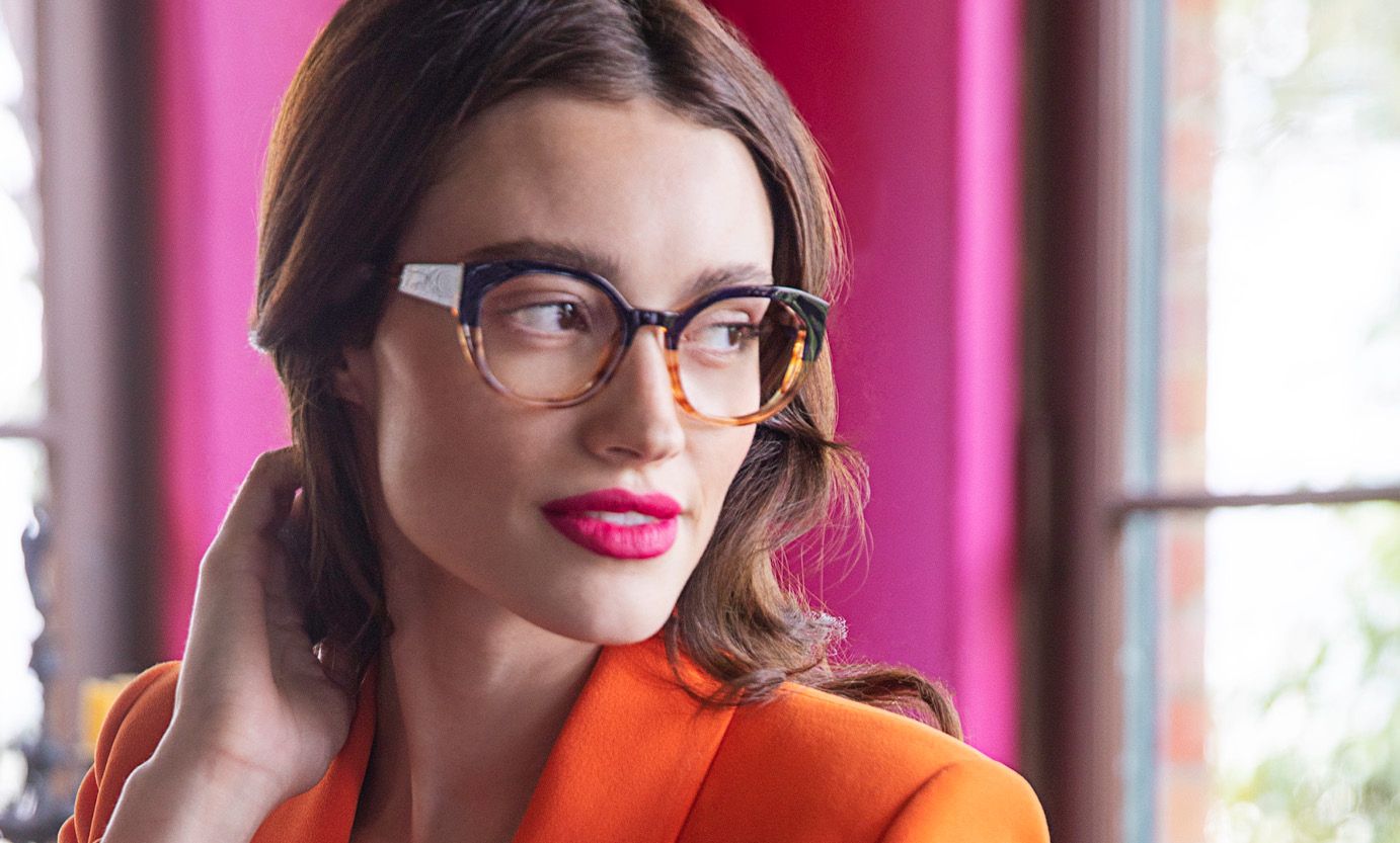 A woman wearing glasses and an orange jacket is standing in front of a pink curtain.