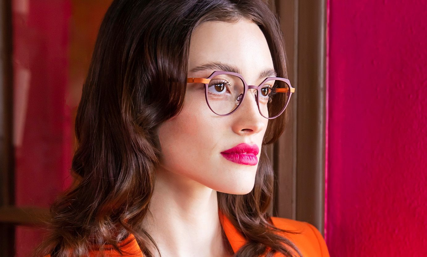 A woman wearing glasses and an orange jacket is looking out of a window.