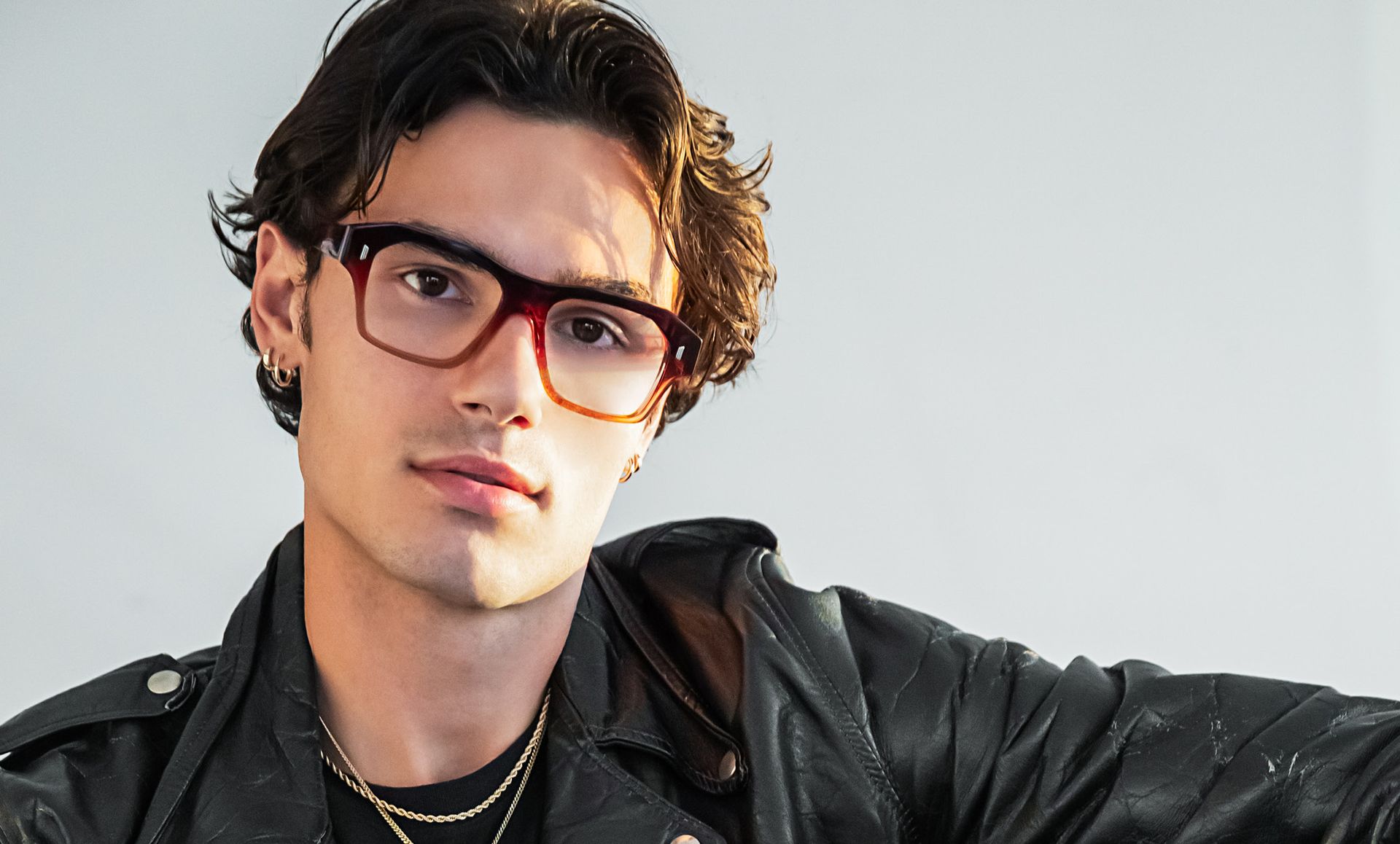 A young man wearing glasses and a leather jacket is looking at the camera.