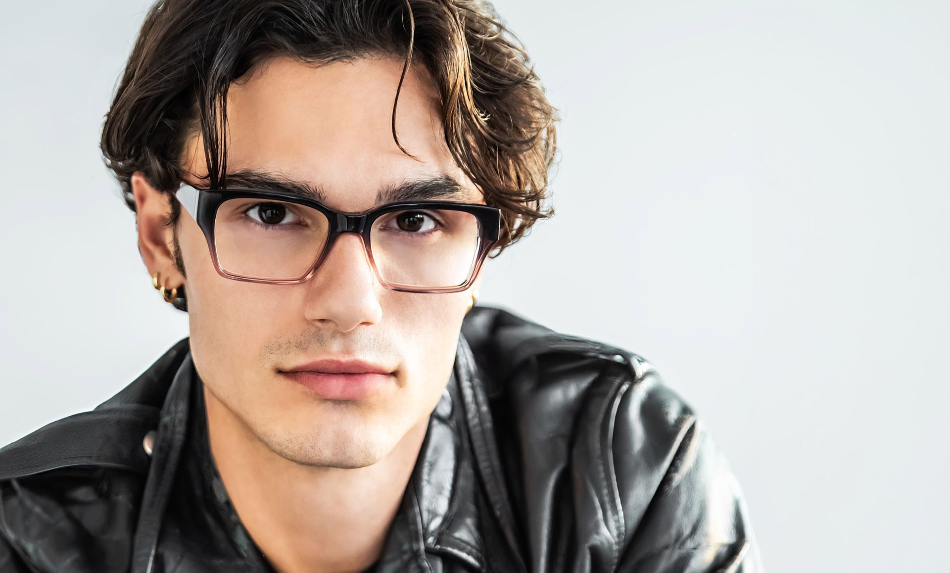 A young man wearing glasses and a leather jacket is looking at the camera.