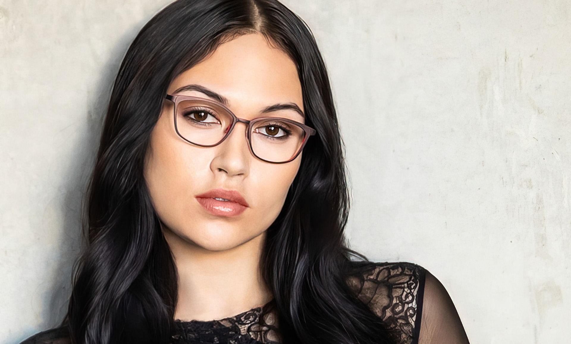 A woman wearing glasses and a black top is standing in front of a wall.