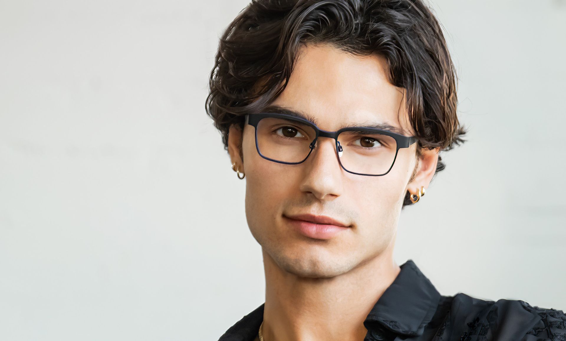 A young man wearing glasses and earrings is looking at the camera.