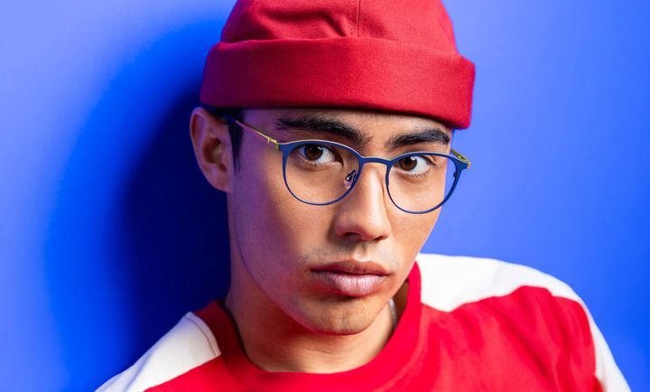 A young man wearing glasses and a red hat is standing in front of a blue wall.