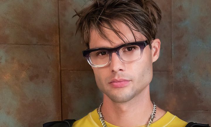 A young man wearing glasses and a yellow shirt is standing in front of a wall.