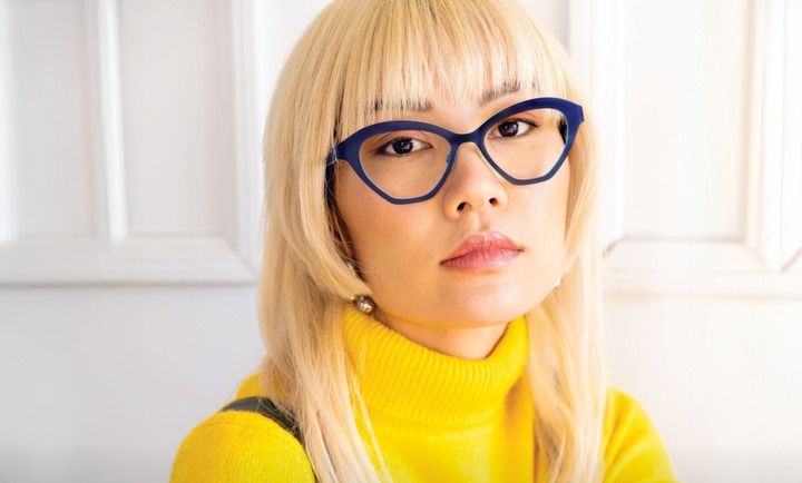 A woman wearing glasses and a yellow sweater is looking at the camera.