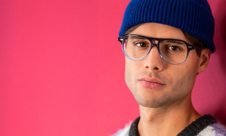 A man wearing glasses and a blue beanie is standing in front of a pink wall.