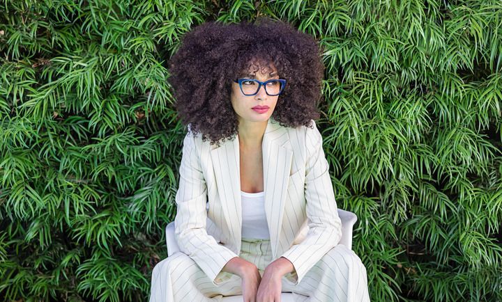 A woman with curly hair and glasses is sitting in front of a bush.