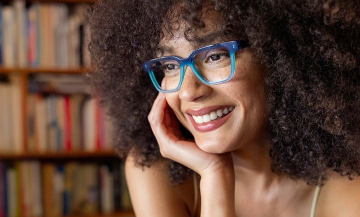 A woman with curly hair and glasses is smiling in front of a bookshelf.