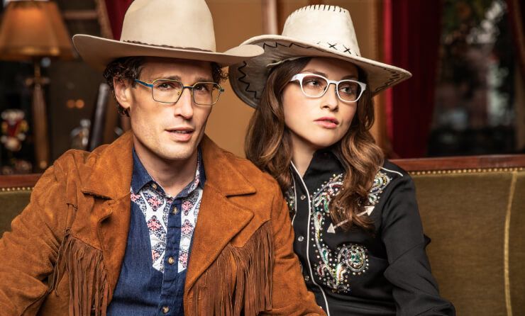 A man and a woman wearing cowboy hats and glasses are sitting on a couch.