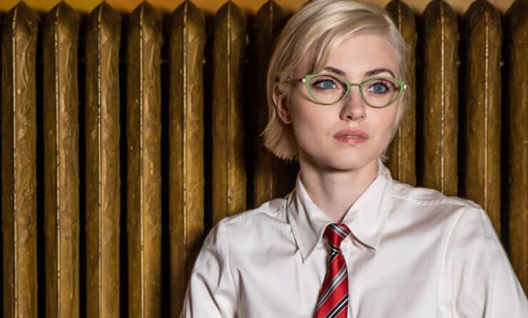 A woman wearing glasses and a tie is standing in front of a radiator.