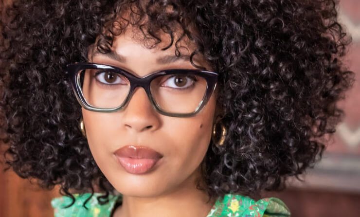 A woman with curly hair and glasses is wearing a green dress.