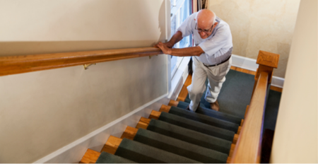 Elderly person ascending a carpeted staircase, using a handrail for support.