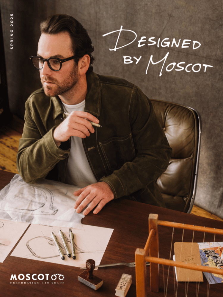 Zack Moscot is wearing glasses and is sitting at a desk with a drawing on it.