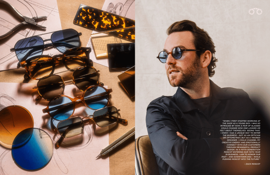 Zack Moscot wearing sunglasses is sitting next to a table with sunglasses on it.